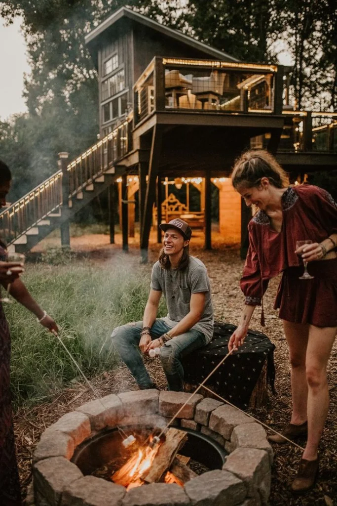 Trois personnes souriantes rôtissent des marshmallows au-dessus d'un feu de camp en pierre, au pied d'une cabane dans les arbres illuminée à la tombée du jour. Une scène conviviale autour d'un logement insolite.