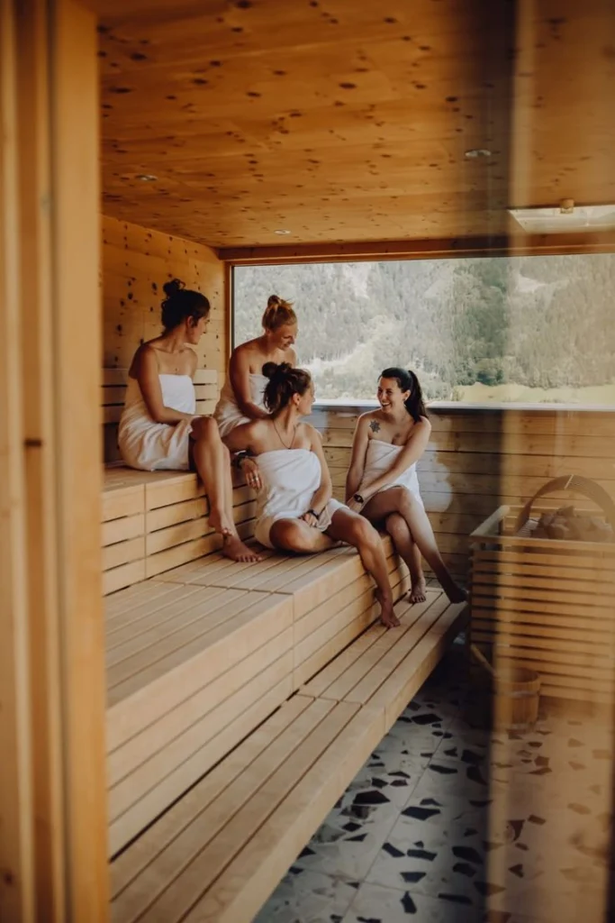 Quatre femmes enveloppées dans des serviettes blanches se détendent et discutent sur les bancs en bois d'un sauna. Une grande fenêtre derrière elles offre une vue sur une vallée verdoyante et des montagnes.