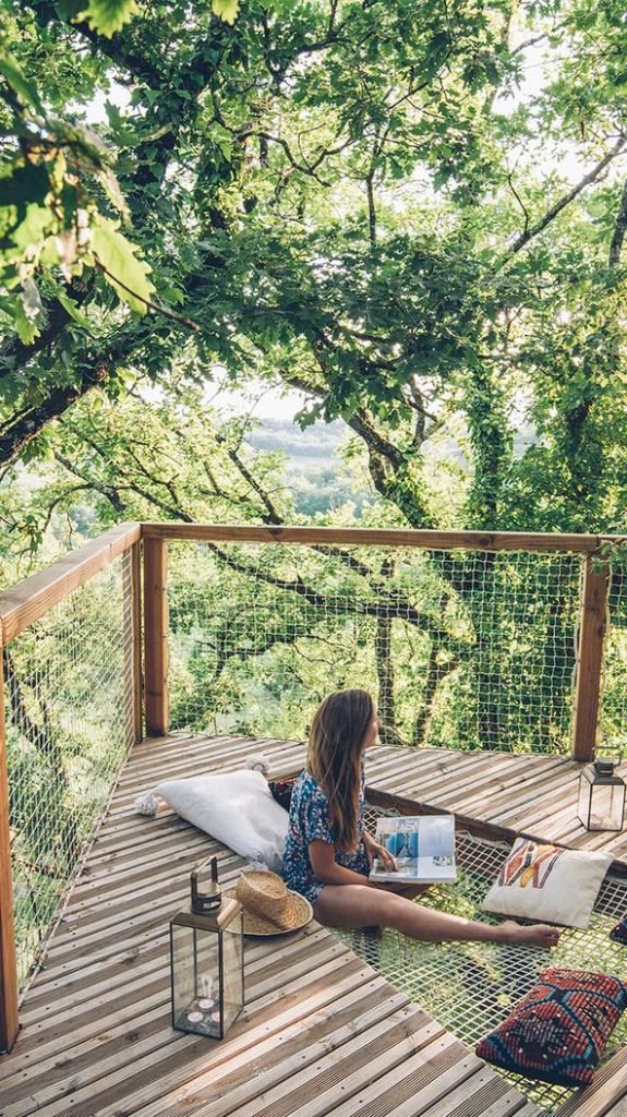 Une femme assise sur un filet de détente intégré à la terrasse en bois d'une cabane dans les arbres, lisant un livre au milieu de la nature verdoyante. Un chapeau de paille et une lanterne sont posés à côté d'elle.