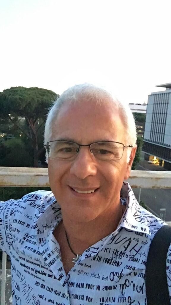 Un homme souriant, aux cheveux poivre et sel, portant des lunettes et une chemise à motifs, prend un selfie en extérieur avec des bâtiments en arrière-plan